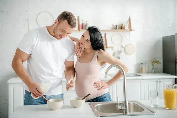 Smiling pregnant woman and man making cereals with milk in kitchen — Stock Photo