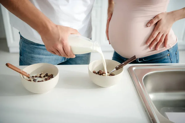 Vista recortada de la pareja embarazada haciendo verter leche en los cereales en la cocina - foto de stock