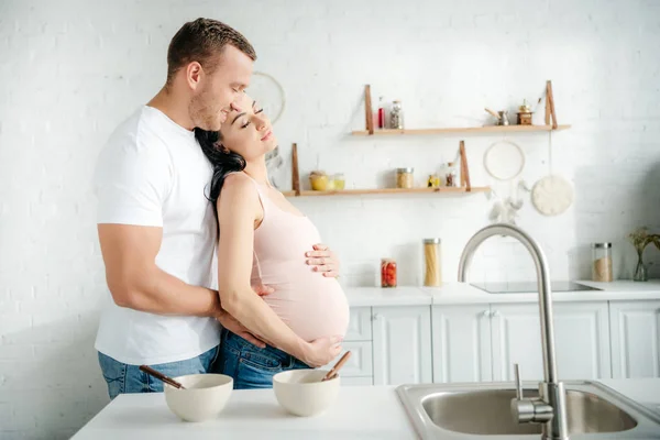 Heureux couple enceinte étreignant dans la cuisine avec des céréales dans des bols — Photo de stock