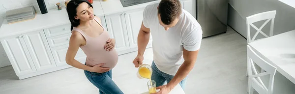 Панорамний знімок красивої усміхненої вагітної дружини на кухні з чоловіком, що поливає апельсиновий сік — стокове фото