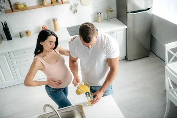 Esposa grávida atraente na cozinha com o marido derramando suco de laranja — Fotografia de Stock