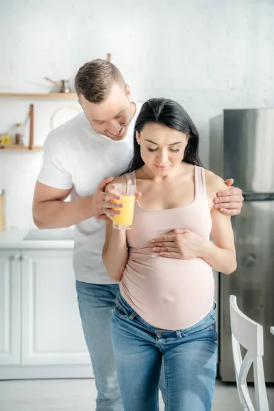 Красива вагітна пара обіймає і тримає апельсиновий сік на кухні — стокове фото