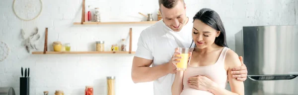 Tiro panorámico de feliz pareja embarazada abrazando y sosteniendo jugo de naranja en la cocina - foto de stock