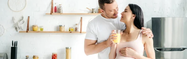 Panoramic shot of happy pregnant couple holding orange juice in kitchen — Stock Photo