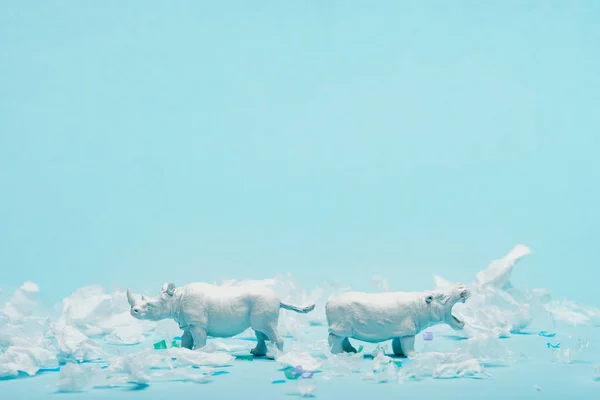 Juguetes blancos de hipopótamo y rinoceronte con basura plástica sobre fondo azul, concepto de bienestar animal - foto de stock