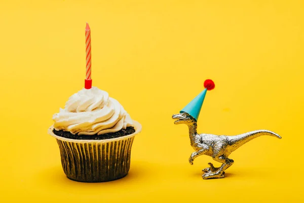 Dinosaurio de juguete en gorra de fiesta y magdalena con vela sobre fondo amarillo - foto de stock