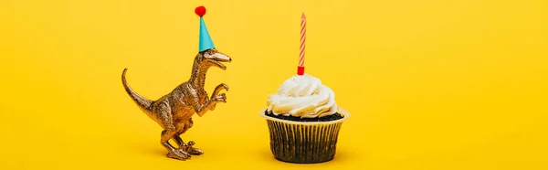 Panoramic shot of toy dinosaur in party cap and cupcake with candle on yellow background — Stock Photo