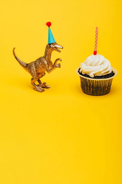 Toy dinosaur in party cap beside cupcake with candle on yellow background — Stock Photo