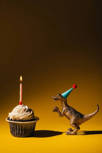 Toy dinosaur in party cap beside cupcake with burning candle on brown background — Stock Photo