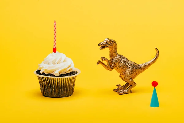 Dinosaurio de juguete junto a la gorra del partido y magdalena con vela sobre fondo amarillo - foto de stock