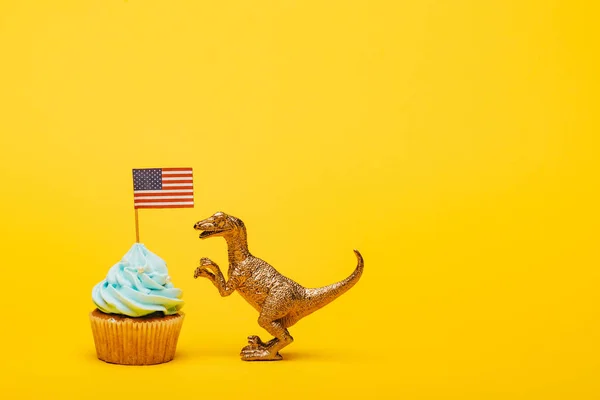 Dinosaurio de juguete junto a magdalena con bandera americana sobre fondo amarillo - foto de stock