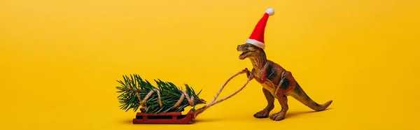 Панорамний знімок іграшкового динозавра в капелюсі Санта з сосною на санях на жовтому фоні — стокове фото