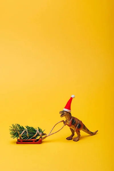 Toy dinosaur in santa hat with fir on sleigh on yellow background — Stock Photo