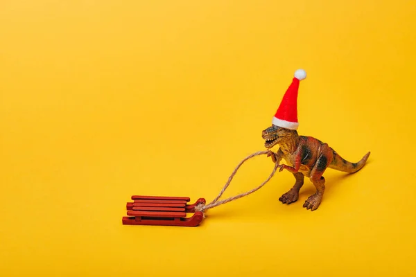 Spielzeug-Dinosaurier mit Weihnachtsmütze und Schlitten auf gelbem Hintergrund — Stockfoto