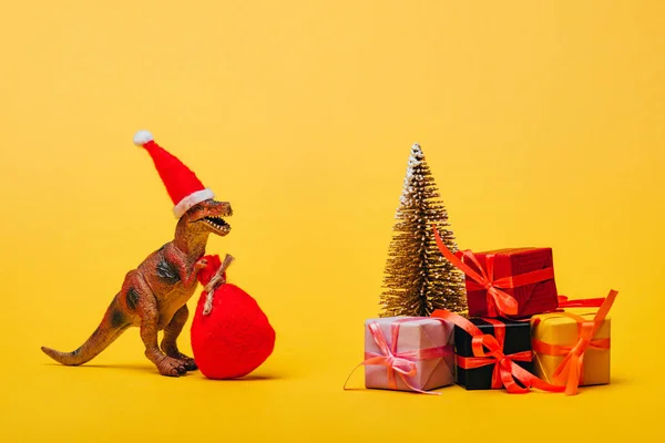 Dinosaurio de juguete en sombrero de santa y saco al lado de pino con regalos sobre fondo amarillo - foto de stock