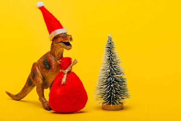 Toy dinosaur in santa hat with sack beside christmas tree on yellow background — Stock Photo