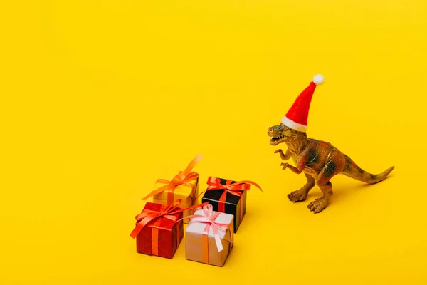 Spielzeug-Dinosaurier mit Weihnachtsmütze neben Geschenken auf gelbem Hintergrund — Stockfoto