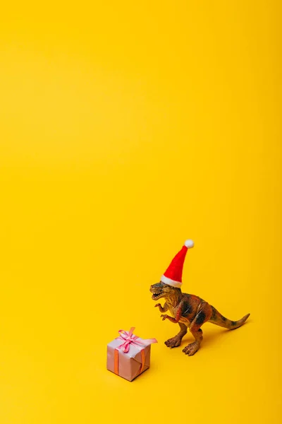 Іграшковий динозавр у капелюсі Санта з подарунком на жовтому тлі — стокове фото