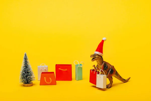 Dinosaurio de juguete en sombrero de santa con bolsas de compras y árbol de Navidad sobre fondo amarillo - foto de stock