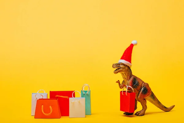 Toy dinosaur in santa hat with shopping bags on yellow background — Stock Photo