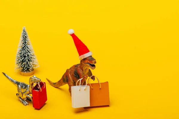 Jouet dinosaures en santa chapeau avec des sacs à provisions et arbre de Noël sur fond jaune — Photo de stock