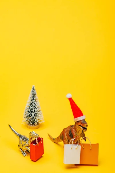 Dinosauri giocattolo in cappello di Babbo Natale con borse della spesa e albero di Natale su sfondo giallo — Foto stock