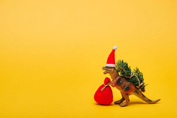 Dinosaurio de juguete en sombrero de santa con saco y abeto sobre fondo amarillo - foto de stock