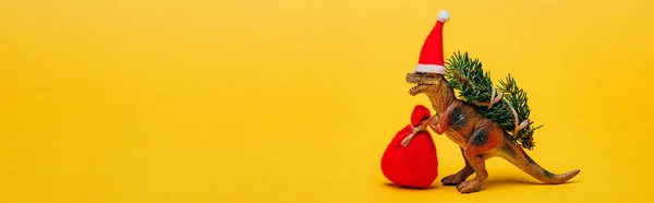 Toy dinosaur in santa hat with sack and fir on yellow background, panoramic shot — Stock Photo