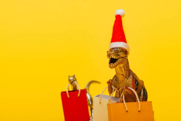 Enfoque selectivo de dinosaurios de juguete en sombrero de santa y bolsas de compras sobre fondo amarillo - foto de stock