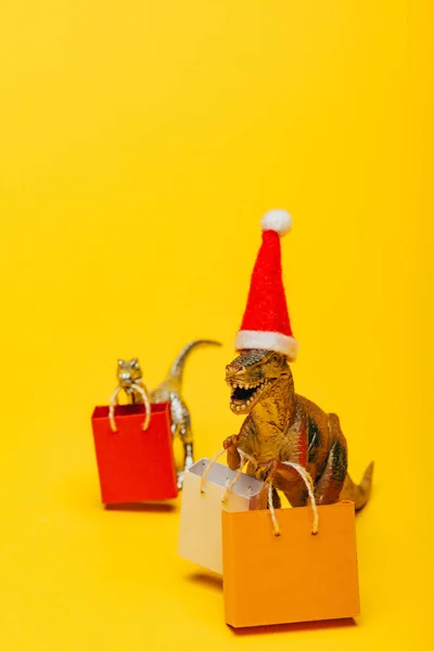 Foco seletivo de dinossauros de brinquedo em chapéu de santa e sacos de compras no fundo amarelo — Fotografia de Stock