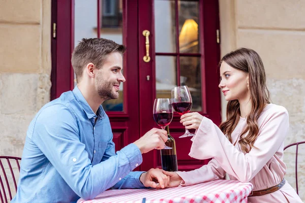 Feliz, pareja elegante sentado en la calle café y tintineo vasos de vino tinto - foto de stock