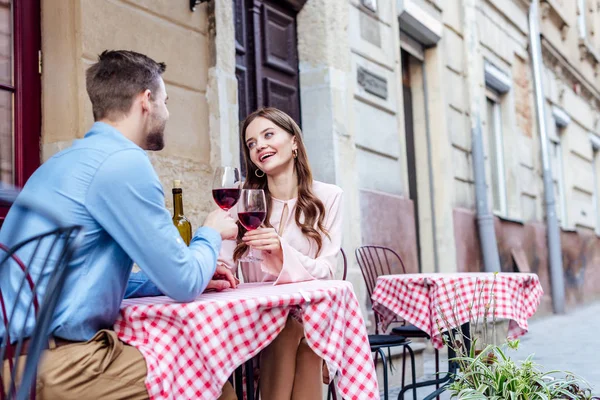 Веселая молодая женщина, звонящая бокалами красного вина с парнем, сидя в уличном кафе — стоковое фото