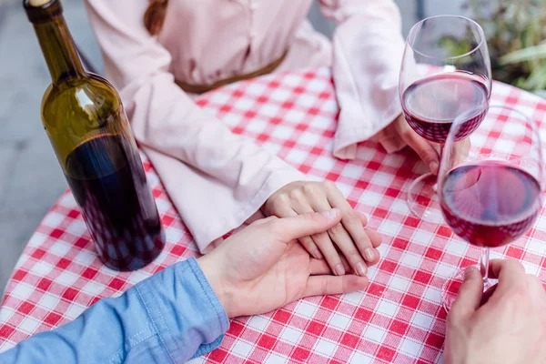 Cropped view of man touching hand of girlfriend while sitting at table with glasses of red wine — Stock Photo