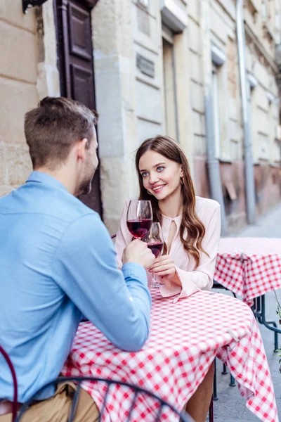 Feliz chica tintineo vasos de vino tinto con novio mientras está sentado en la calle café - foto de stock