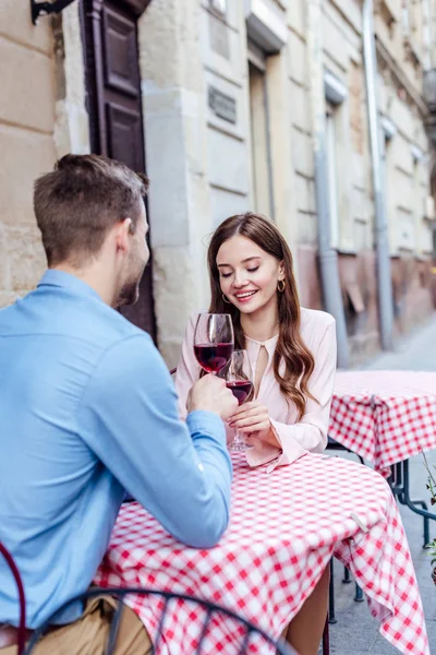 Happy young woman clinking glasses of red wine with boyfriend while sitting in street cafe — Stock Photo