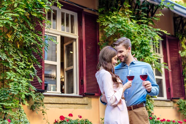 Feliz joven pareja sosteniendo vasos de vino tinto mientras está de pie cerca de la casa cubierta de hiedra verde - foto de stock