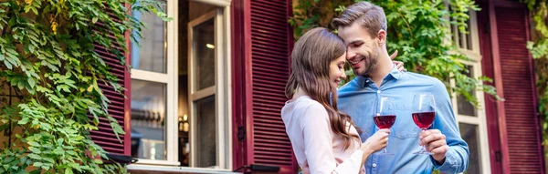 Plan panoramique de couple heureux tenant des verres de vin rouge tout en se tenant près de la maison couverte de lierre vert — Photo de stock