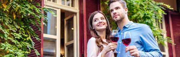 Panoramic shot of happy couple holding glasses of red wine while standing near house covered with green ivy — Stock Photo