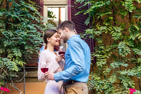 Glückliches Paar, das sich mit geschlossenen Augen umarmt und Gläser Rotwein hält, während es in der Nähe des mit grünem Efeu bedeckten Hauses steht — Stockfoto