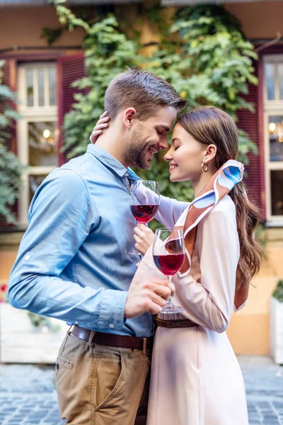 Vista lateral de feliz pareja joven abrazando con los ojos cerrados mientras sostiene vasos de vino tinto en la calle - foto de stock