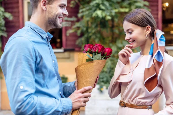 Jovem bonito apresentando buquê de rosas vermelhas para namorada feliz — Fotografia de Stock