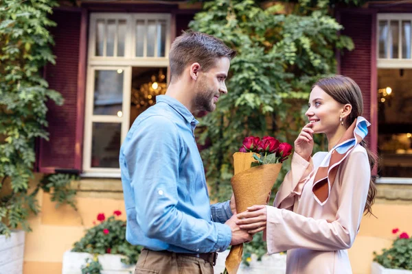 Side view of young man presenting bouquet of roses to happy girlfriend on street — Stock Photo