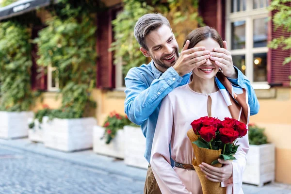 Hombre feliz cerrando los ojos de la novia sonriente sosteniendo ramo de rosas - foto de stock