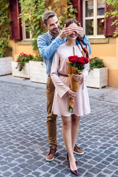 Веселий чоловік закриває очі усміхненої дівчини, що тримає букет троянд. — стокове фото