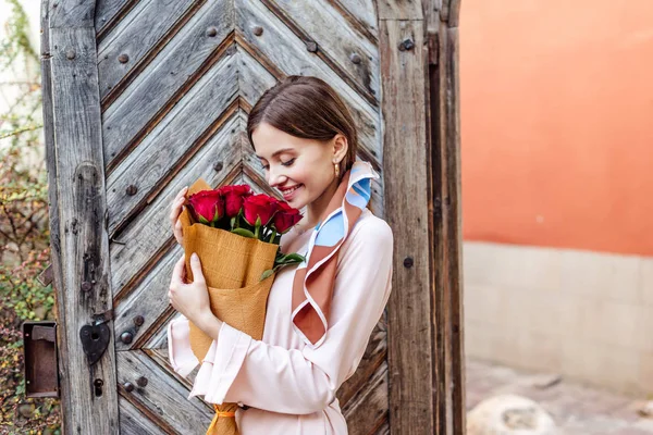 Panoramic shot of happy girl holding bouquet of red roses while standing near old wooden door — Stock Photo