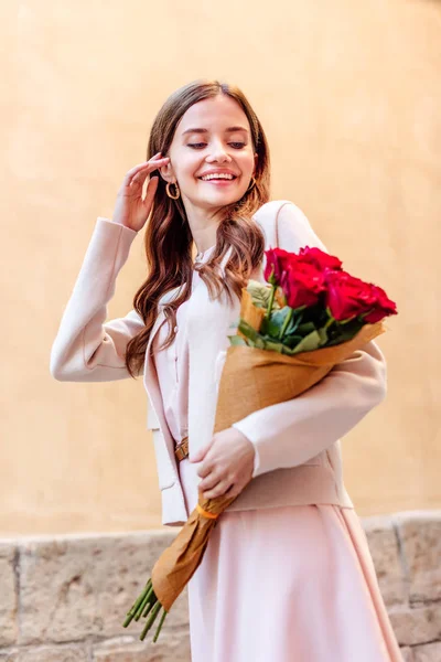 Happy girl holding bouquet of red roses and touching hair while walking on street — Stock Photo
