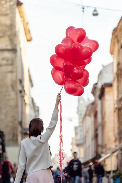 Visão traseira da jovem mulher segurando pacote de balões em forma de coração vermelho na rua — Fotografia de Stock