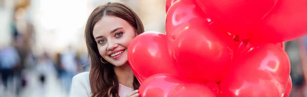 Foto panoramica di bella ragazza sorridente alla macchina fotografica mentre tiene in mano fascio di palloncini rossi a forma di cuore — Foto stock