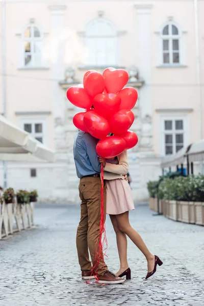 Feliz jovem casal abraçando enquanto se esconde atrás de balões em forma de coração vermelho na praça da cidade — Fotografia de Stock