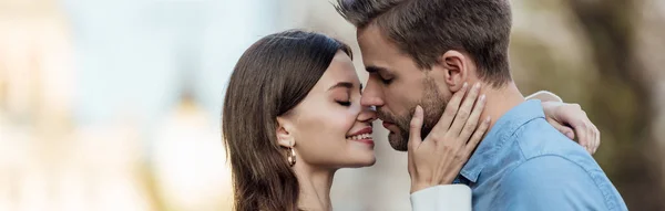 Tiro panorámico de chica feliz con los ojos cerrados besando novio guapo en la calle - foto de stock